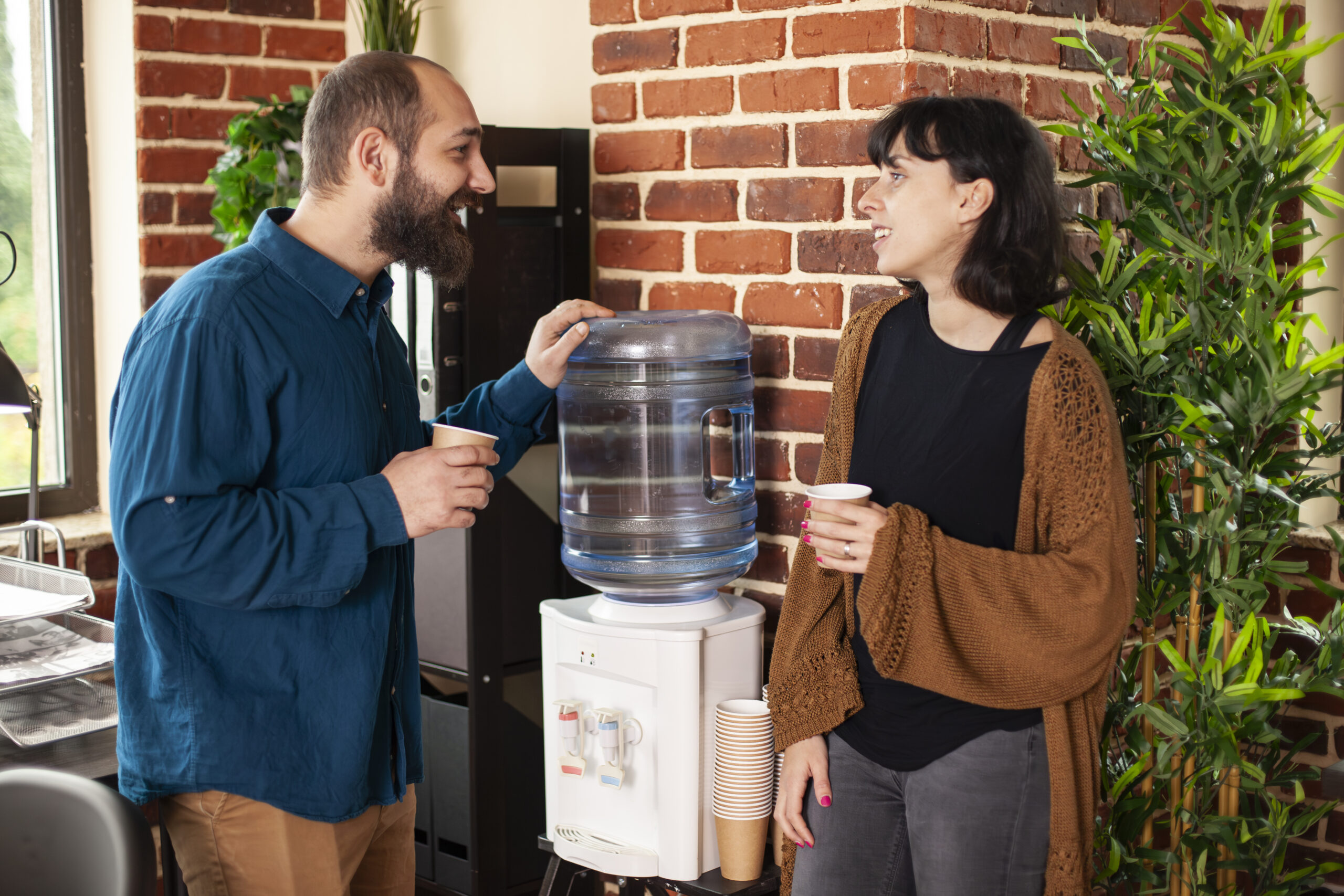 Bearded man and female colleague hold paper cups standing near brick wall and exchanging business project ideas. Businesswoman and male coworker stand next to water dispenser taking break at office.